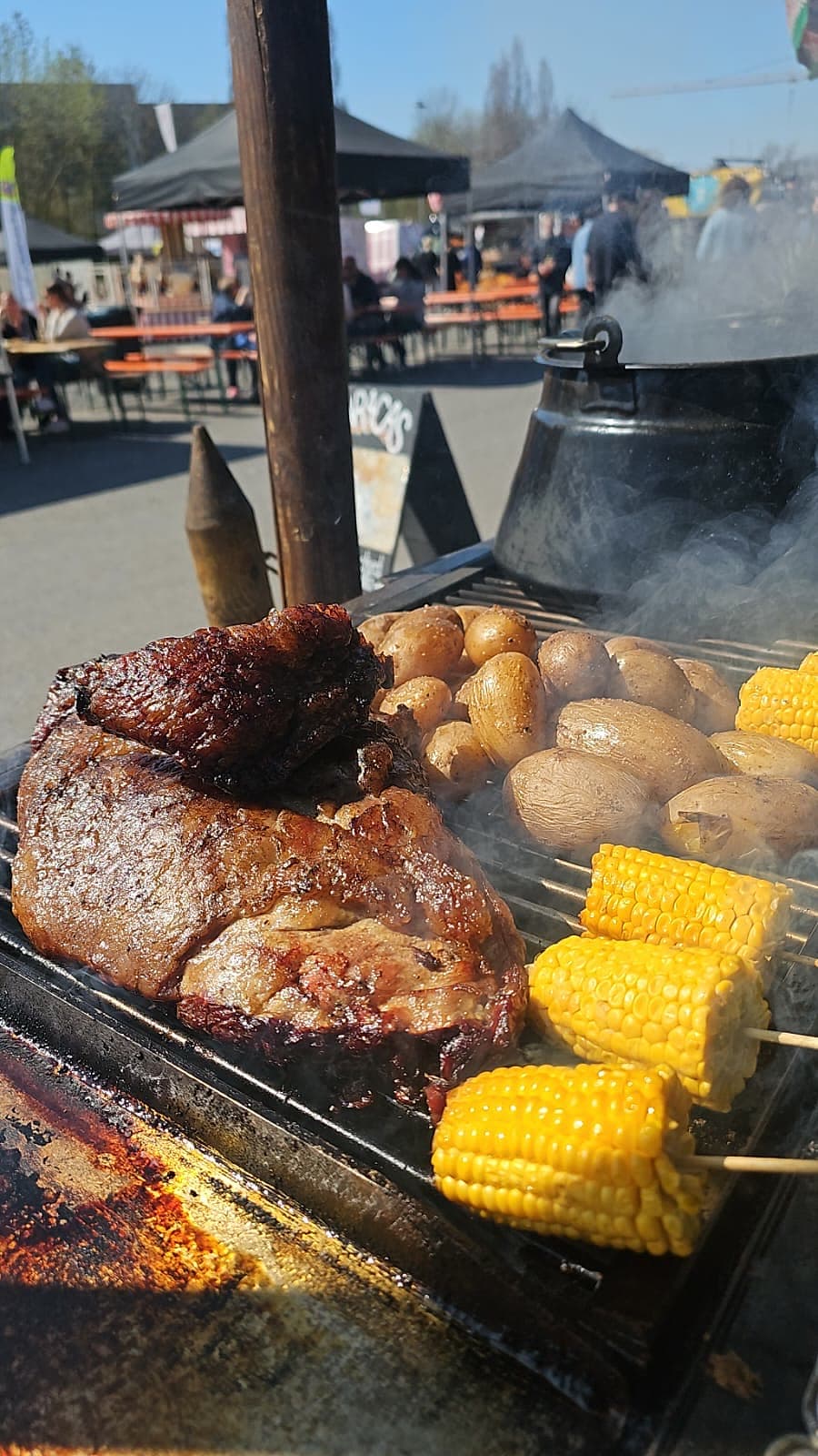 Smoked Beef, Papas und Mais am Spieß auf der Parrilla — Rauch zieht über das Fleisch, gusseiserner Topf im Hintergrund