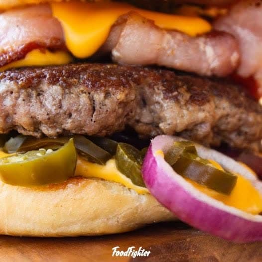 Close-up des Signature-Burgers "Den will ich haben" — Bacon, geschmolzener Cheddar, Jalapeños, rote Zwiebel auf fluffiger Brioche