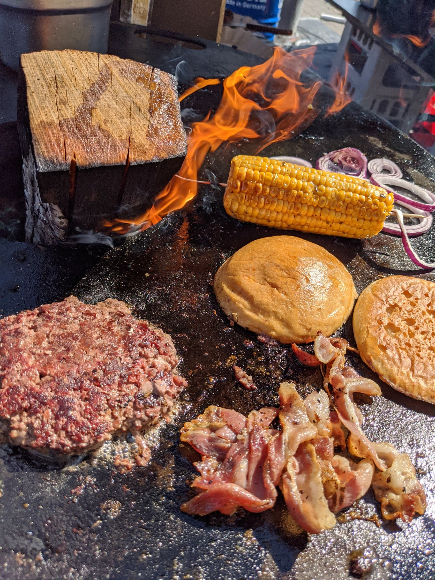Plancha über offenem Feuer mit Holzscheit, rohem Smash-Patty, Bacon, Maiskolben und fluffiger Brioche