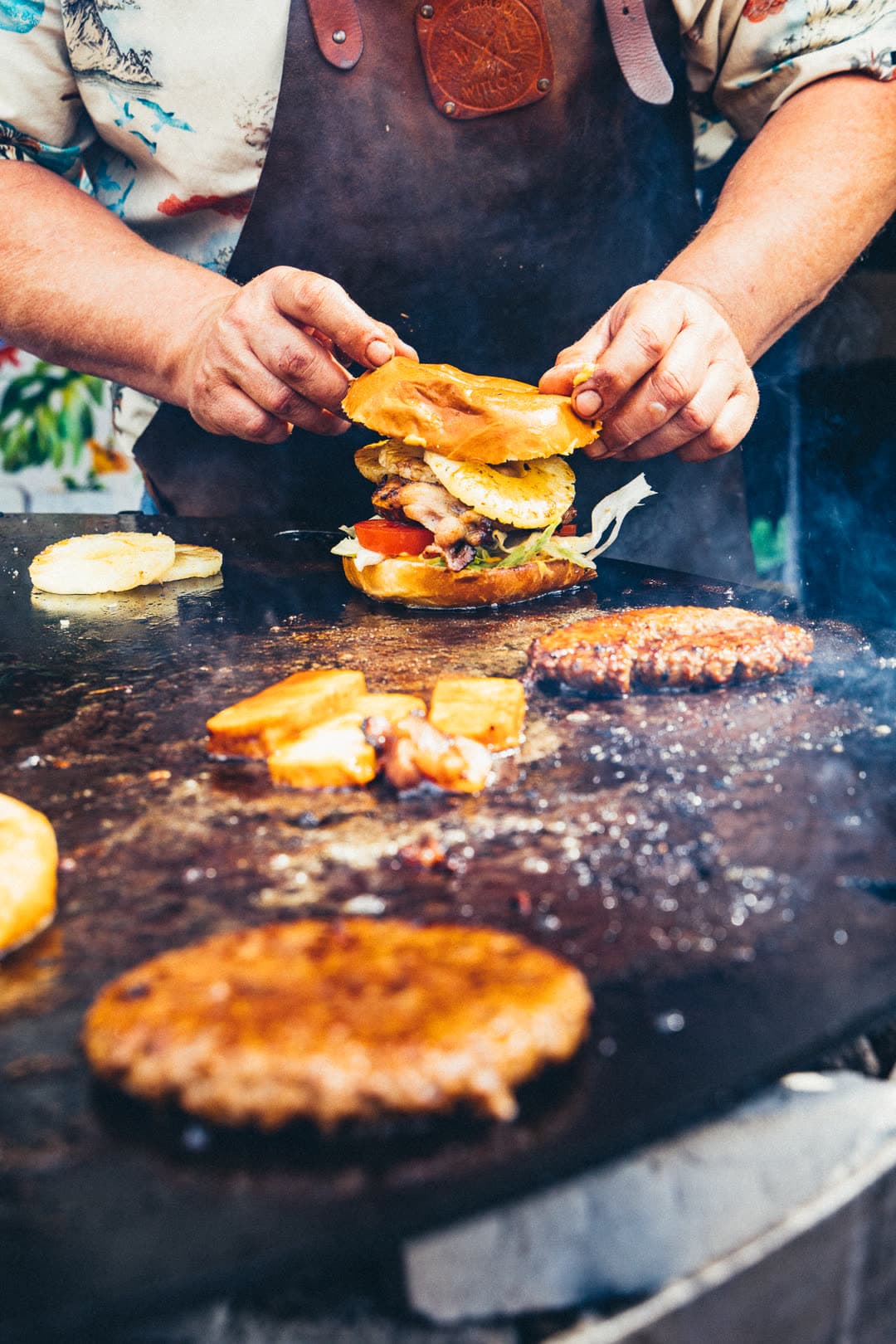 Plancha-Detail mit Patties, Bacon und gegrillten Ananasscheiben über offener Flamme