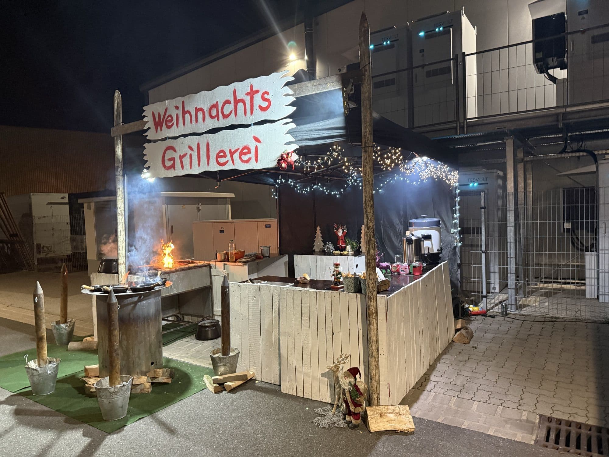Die FoodFighter Weihnachtsgrillerei am Abend: Holzstand mit Lichterketten, Feuertonne und Schild „Weihnachts Grillerei“